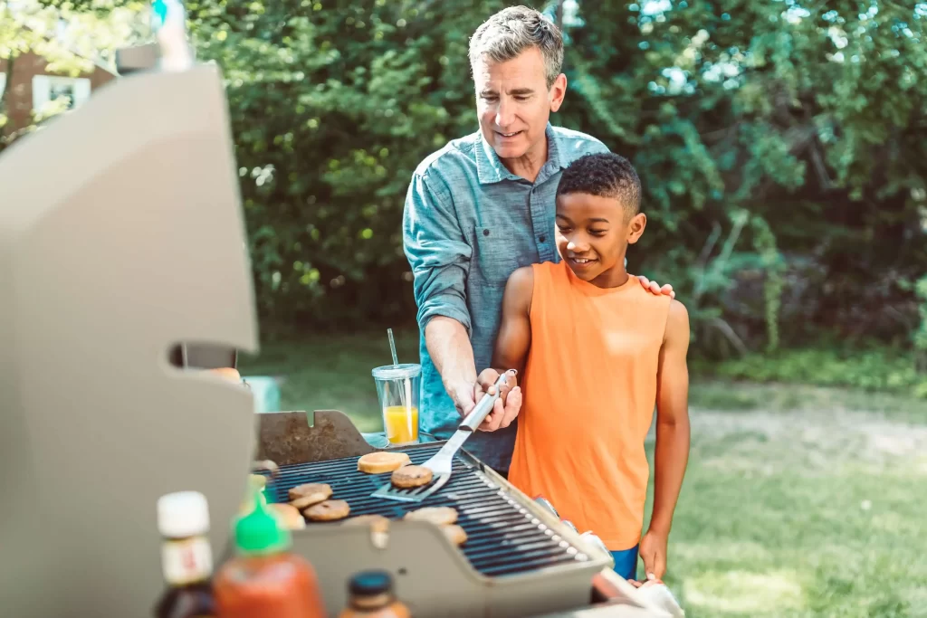 Top 5 Outdoor Amenities For HOAs & Condos 4 older white man in blue shirt shows young black boy in an orange top how to grill a burger. HOA outdoor amenities.