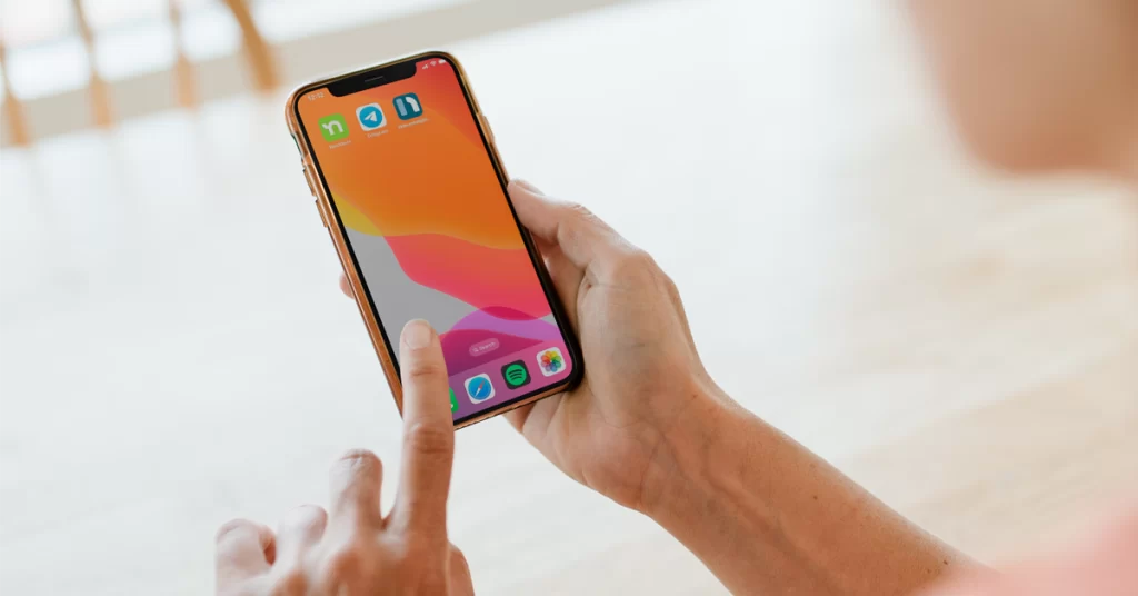 The 3 Best HOA Communication Apps To Easily Engage With Residents 3 a hand holding a smartphone with an orange screensaver with 3 hoa communication apps: nextdoor, telegram and neigbrs by vinteum. It also has spotify, safari and image gallery, with a finger pointing at a screen