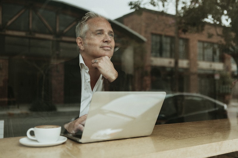 5 Creative Ideas for your HOA Newsletter + Free Templates 1 Older man with white hair in a jacket and white shirt looking reflective looking out of the window with a laptop and coffee next to him, thinking about HOA Newsletter