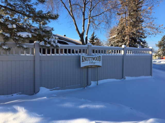 Customer Success Stories: Knottwood Village & Neigbrs by Vinteum 1 Snowy light brown fence with a sign that says Knottwood Village. There is snow piled up in front and with trees behind